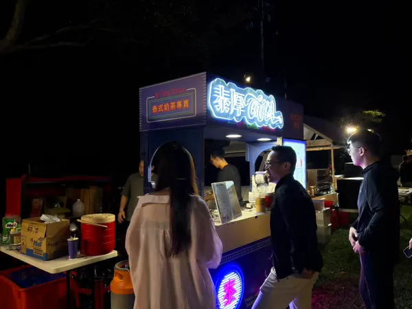 攤車前賓客邊聊天邊等飲料,招牌與泰字燈箱亮著
